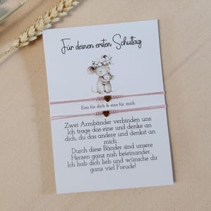 Könnte beinhalten: Eine weiße Karte mit deutschem Text und einer Zeichnung einer Mutter und Babymaus. Zwei rosa Armbänder sind an der Karte befestigt. Der Text auf der Karte lautet "Für deinen ersten Schultag".
