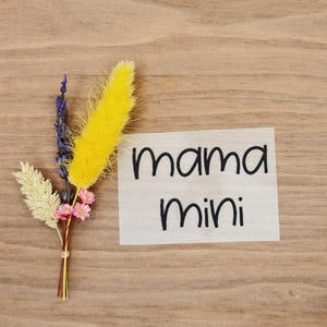 Könnte beinhalten: Ein kleiner Strauß Trockenblumen mit gelben, lila und rosa Akzenten, zusammengebunden mit einem dünnen goldenen Faden. Ein helles Holzschild mit den Worten "mama mini" in schwarzer Schrift befindet sich neben dem Strauß.