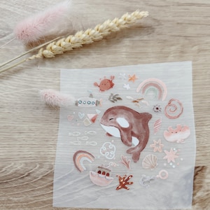 Könnte beinhalten: Ein Bogen mit Aufklebern zum Thema Ozean mit einem braunen und weißen Orca, Regenbögen, Muscheln und anderen Meereskreaturen. Die Aufkleber sind in Rosa-, Orange- und Brauntönen gehalten. Der Bogen liegt auf einer Holzoberfläche mit getrockneten Blumen.