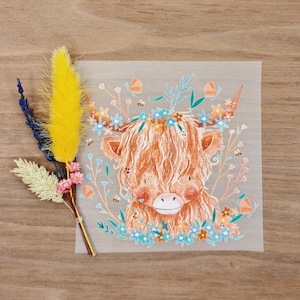 Könnte beinhalten: Quadratischer Druck mit einer Illustration einer Highland-Kuh, geschmückt mit einem Blumenkranz und umgeben von Blumen, Schmetterlingen und Bienen. Die Kuh hat orangefarbenes Fell, mit blauen, orangefarbenen und grünen floralen Elementen. Ein Strauß getrockneter Blumen in Gelb, Blau und Rosa befindet sich links.