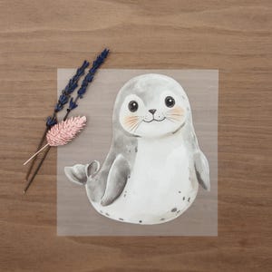 Könnte beinhalten: Aquarellillustration eines grauen Seehunds mit weißem Bauch und rosigen Wangen. Der Seehund lächelt und hat schwarze Augen. Ein Zweig getrockneten Lavendels und eine rosa Blume befinden sich links vom Seehund.