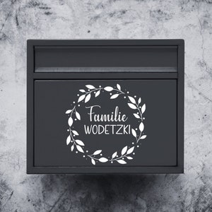 Puede incluir: Un buz&oacute;n negro con una corona blanca y el texto "Familie WODETZKI" en la parte delantera.