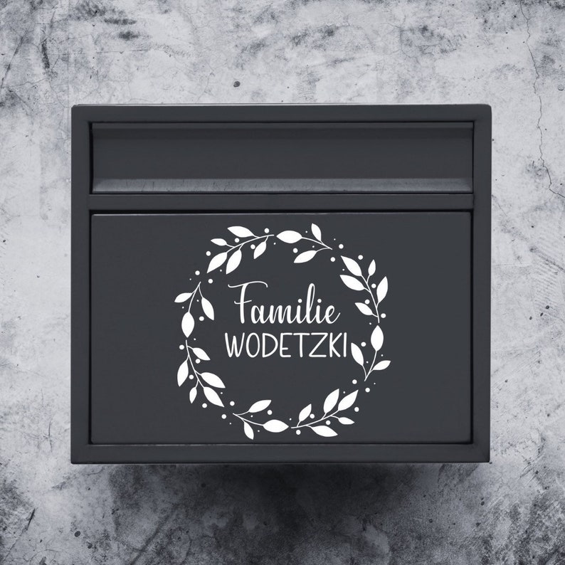 Puede incluir: Un buz&oacute;n negro con una corona blanca y el texto "Familie WODETZKI" en la parte delantera.