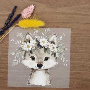 Könnte beinhalten: Aquarell-Illustration eines Wolfes mit einem Blumenkranz aus weißen Gänseblümchen und grünen Blättern. Der Wolf hat graues und weißes Fell, mit großen, dunklen Augen und einer kleinen schwarzen Nase. Das Bild hat einen transparenten Hintergrund.