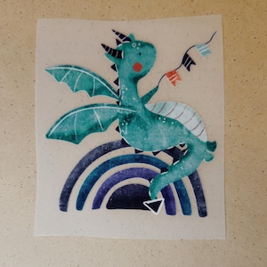 Könnte beinhalten: Eine skurrile Illustration eines türkisfarbenen Drachen mit roter Wange, der ein Banner mit Flaggen hält. Der Drache sitzt auf einem blauen und lila Regenbogen. Der Drache hat Flügel und einen weißen Bauch.