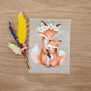Könnte beinhalten: Aquarellillustration von zwei Füchsen, einer umarmt den anderen. Der größere Fuchs trägt einen Blumenkranz. Die Füchse sind in Orange- und Weißtönen gehalten. Links ein kleiner Strauß getrockneter Blumen in Gelb, Blau und Rosa.