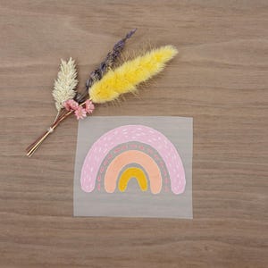Könnte beinhalten: Ein farbenfroher Regenbogen-Aufkleber mit einem rosa, orangefarbenen und gelben Design. Der Aufkleber befindet sich auf einer transparenten Folie und neben einem kleinen Strauß Trockenblumen mit gelben, weißen und rosa Akzenten. Der Hintergrund ist Holz.
