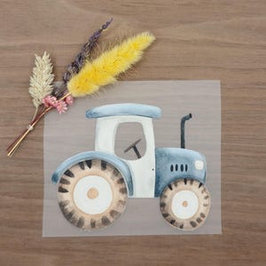 Könnte beinhalten: Aquarellillustration eines blau-weißen Traktors mit braunen Rädern. Der Traktor hat einen schwarzen Schornstein und ein Lenkrad. Ein kleiner Strauß Trockenblumen in Gelb-, Rosa- und Beigetönen befindet sich links vom Traktor.