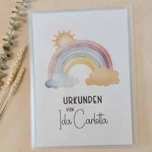 Könnte beinhalten: Ein klarer Kunststoff-Dokumentenhalter mit einem Aquarelldruck eines Regenbogens, einer Sonne und Wolken. Der Text "URKUNDEN von Ida Carlotta" ist unter dem Regenbogen aufgedruckt. Der Halter ist von Zweigen umgeben.