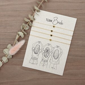 Könnte beinhalten: Eine Karte mit dem Text "Team Bride" und einer Zeichnung von drei Frauen, die mit ihren Händen Herzformen bilden. Vier goldene Herz-Charm-Armbänder sind an der Karte befestigt. Ein Zweig Eukalyptus und eine rosa Trockenblume befinden sich links.