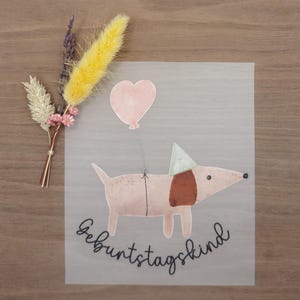 Könnte beinhalten: Hellrosa Illustration eines Hundes mit Partyhut und herzförmigem Ballon. Das deutsche Wort "Geburtstagskind" ist darunter geschrieben. Getrocknete Blumen in Gelb, Weiß und Rosa befinden sich in der oberen linken Ecke.