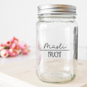 Könnte beinhalten: Ein klares Glasgefäß mit einem silbernen Deckel. Das Gefäß hat ein schwarzes Etikett mit der Aufschrift "Müsli FRUCHT".
