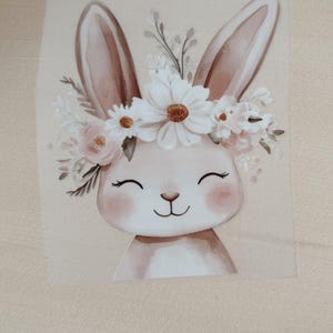 Könnte beinhalten: Illustration eines lächelnden Hasen mit Blumenkranz. Der Hase hat lange, aufrechte Ohren und eine hellbraune Farbe. Der Blumenkranz zeigt weiße Gänseblümchen und rosa Blüten. Der Hintergrund ist hellbeige.