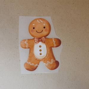 Könnte beinhalten: Ein Aufkleber mit einem lächelnden Lebkuchenmann, einer Fliege und drei Knöpfen. Der Aufkleber ist hellbraun mit weißen Zuckergussdetails und befindet sich auf einem weißen Hintergrund.