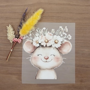 Könnte beinhalten: Eine bezaubernde Illustration einer lächelnden Maus mit einem Blumenkranz. Die Maus ist in sanften, neutralen Tönen mit rosigen Wangen und einer kleinen rosa Nase dargestellt. Links befindet sich ein kleiner Strauß aus getrockneten Blumen in Gelb, Rosa und Beige.