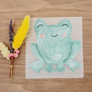 Könnte beinhalten: Eine Illustration im Aquarellstil einer lächelnden grünen Frosches mit rosa Wangen. Der Frosch befindet sich auf einem quadratischen, durchscheinenden Hintergrund. Links vom Frosch befindet sich ein kleiner Strauß aus getrockneten Blumen in Gelb, Lila und Rosa.