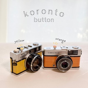 May include: Two vintage Olympus Trip 35 cameras, one yellow and one orange. The text "koronto button" is above the cameras. The yellow camera has the text "Olympus 35 DC" on it. The orange camera has the text "Olympus Trip 35" on it.