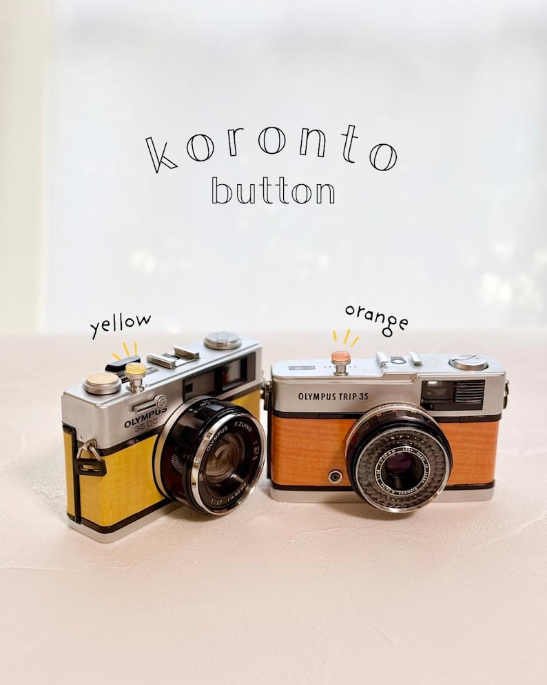 May include: Two vintage Olympus Trip 35 cameras, one yellow and one orange. The text "koronto button" is above the cameras. The yellow camera has the text "Olympus 35 DC" on it. The orange camera has the text "Olympus Trip 35" on it.