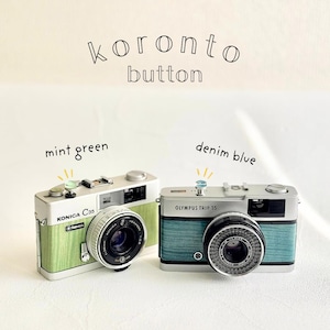 May include: Two vintage cameras, a Konica C35 in mint green and an Olympus Trip 35 in denim blue, are displayed. Each camera has a colored button on top. The text "koronto button" is above the cameras.