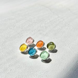 May include: Seven colourful stud earrings. Each earring has a round, translucent gem in shades of yellow, pink, orange, blue, green, and a lighter green. The gems are set in silver-coloured metal.