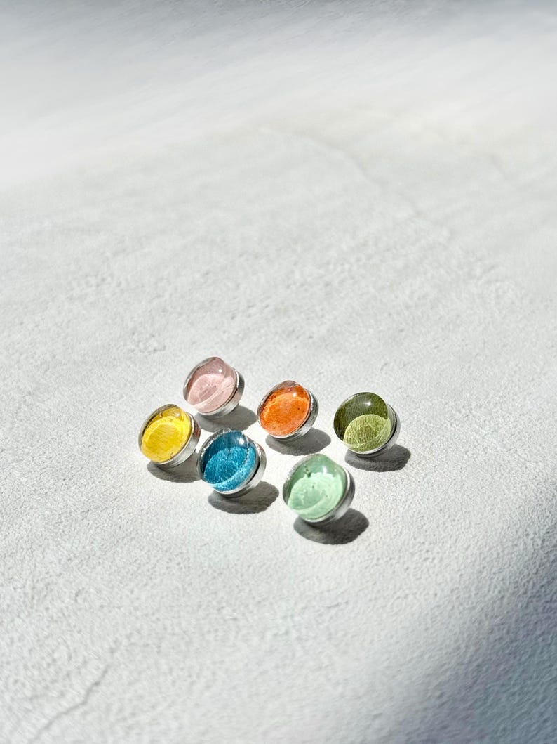 May include: Seven colourful stud earrings. Each earring has a round, translucent gem in shades of yellow, pink, orange, blue, green, and a lighter green. The gems are set in silver-coloured metal.