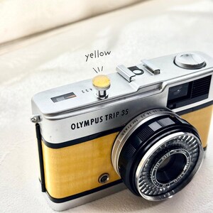 May include: A vintage Olympus Trip 35 camera with a yellow body and silver detailing. The camera lens is black with silver detailing. The text "OLYMPUS TRIP 35" is printed on the front. The word "yellow" is written above the camera's button.