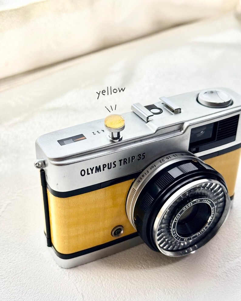 May include: A vintage Olympus Trip 35 camera with a yellow body and silver detailing. The camera lens is black with silver detailing. The text "OLYMPUS TRIP 35" is printed on the front. The word "yellow" is written above the camera's button.