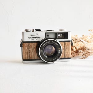 Könnte beinhalten: Olympus 35 DC Kamera mit silbernem Gehäuse, Holzmaserung und schwarzem Objektiv. Das Objektiv ist mit "Olympus F. Zuiko 1:1.7 f=40mm" beschriftet. Die Kamera steht auf einer weißen Oberfläche.