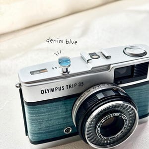 May include: An Olympus Trip 35 camera with a denim blue finish. The camera has a black lens and the text "OLYMPUS TRIP 35" is visible. The image also includes the text "denim blue".