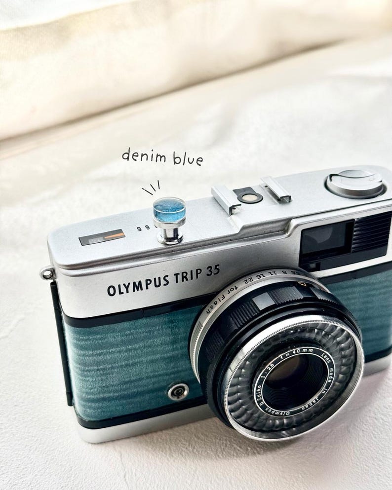 May include: An Olympus Trip 35 camera with a denim blue finish. The camera has a black lens and the text "OLYMPUS TRIP 35" is visible. The image also includes the text "denim blue".