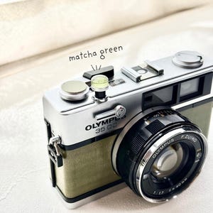 May include: A vintage Olympus 35 DC camera with a matcha green body and silver detailing. The camera has a black lens with the text "Olympus F.Zuiko 1:1.7 f=40mm". The words "matcha green" are written above the camera.