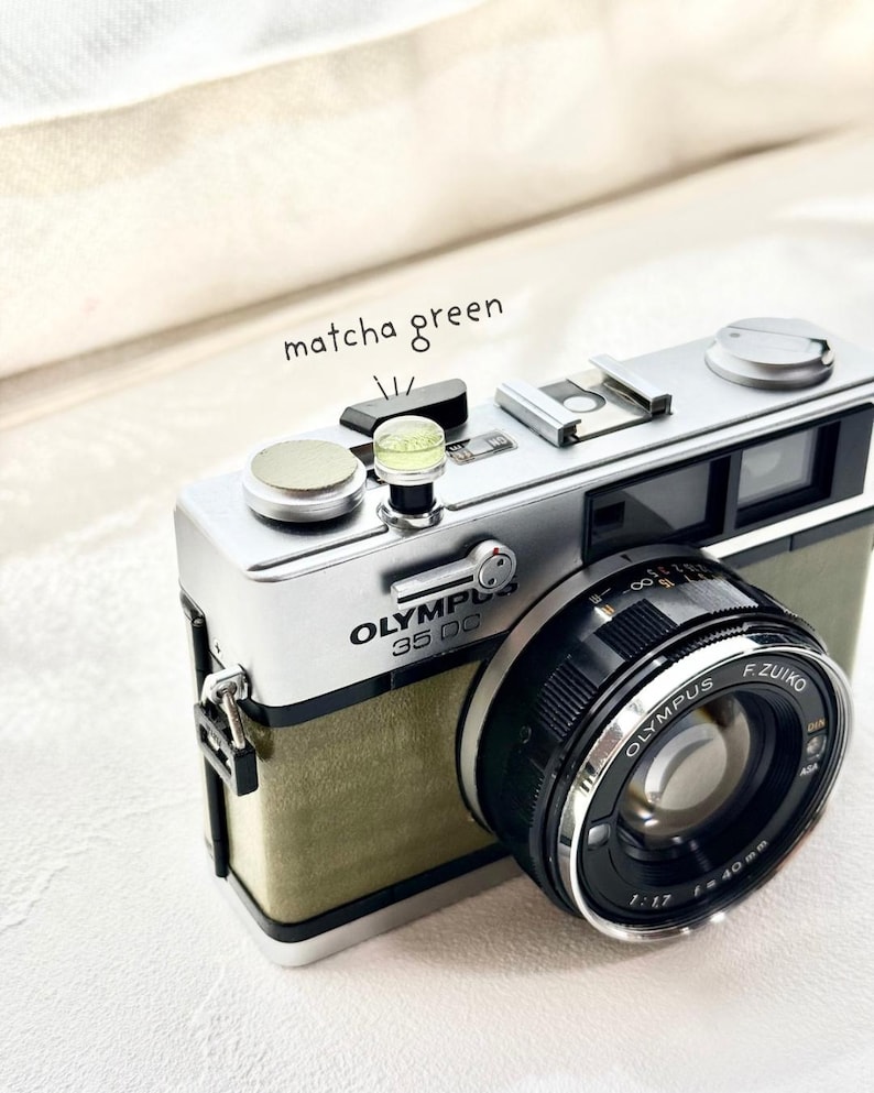 May include: A vintage Olympus 35 DC camera with a matcha green body and silver detailing. The camera has a black lens with the text "Olympus F.Zuiko 1:1.7 f=40mm". The words "matcha green" are written above the camera.
