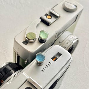 May include: Close-up of two vintage silver cameras with black lenses and colourful accents. One camera has the text "Yashica C35" and the other has the number "334166" printed on it. The cameras are on a white surface.