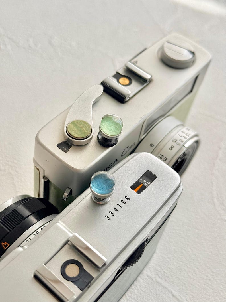 May include: Close-up of two vintage silver cameras with black lenses and colourful accents. One camera has the text "Yashica C35" and the other has the number "334166" printed on it. The cameras are on a white surface.