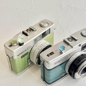May include: Two vintage film cameras, a Konica C35 and an Olympus Trip 35, are shown. The Konica has a green textured finish, while the Olympus has a blue finish. Both cameras have silver accents and lenses, evoking a retro style.