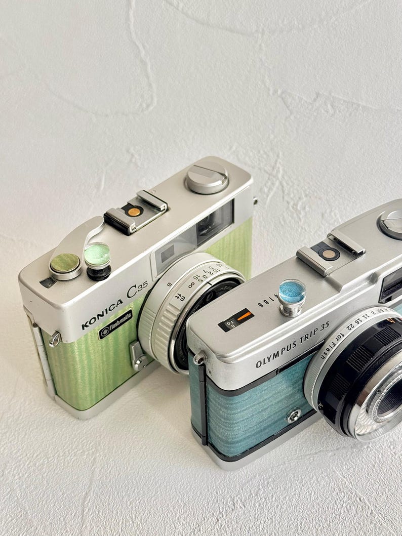 May include: Two vintage film cameras, a Konica C35 and an Olympus Trip 35, are shown. The Konica has a green textured finish, while the Olympus has a blue finish. Both cameras have silver accents and lenses, evoking a retro style.