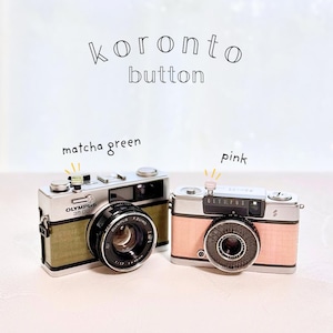 May include: Two vintage-style Olympus 35 DC cameras. One is matcha green, the other is pink. The cameras have black lenses and silver accents. The text "koronto button" is above the cameras. The word "Olympus" is printed on the cameras.