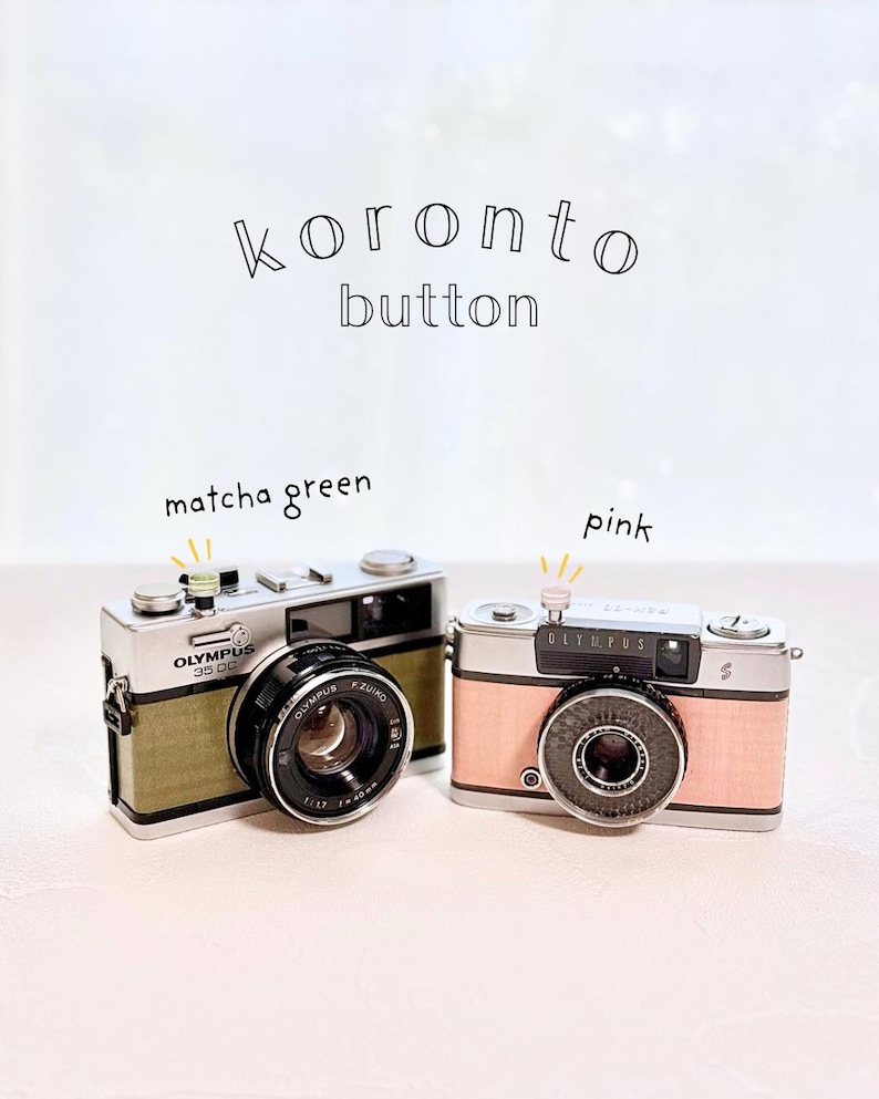 May include: Two vintage-style Olympus 35 DC cameras. One is matcha green, the other is pink. The cameras have black lenses and silver accents. The text "koronto button" is above the cameras. The word "Olympus" is printed on the cameras.
