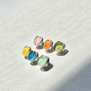 May include: A selection of colourful stud earrings. Each earring has a silver-toned metal base and a translucent, jewel-toned centre in shades of pink, orange, green, blue, and yellow. The earrings are arranged on a light grey surface.