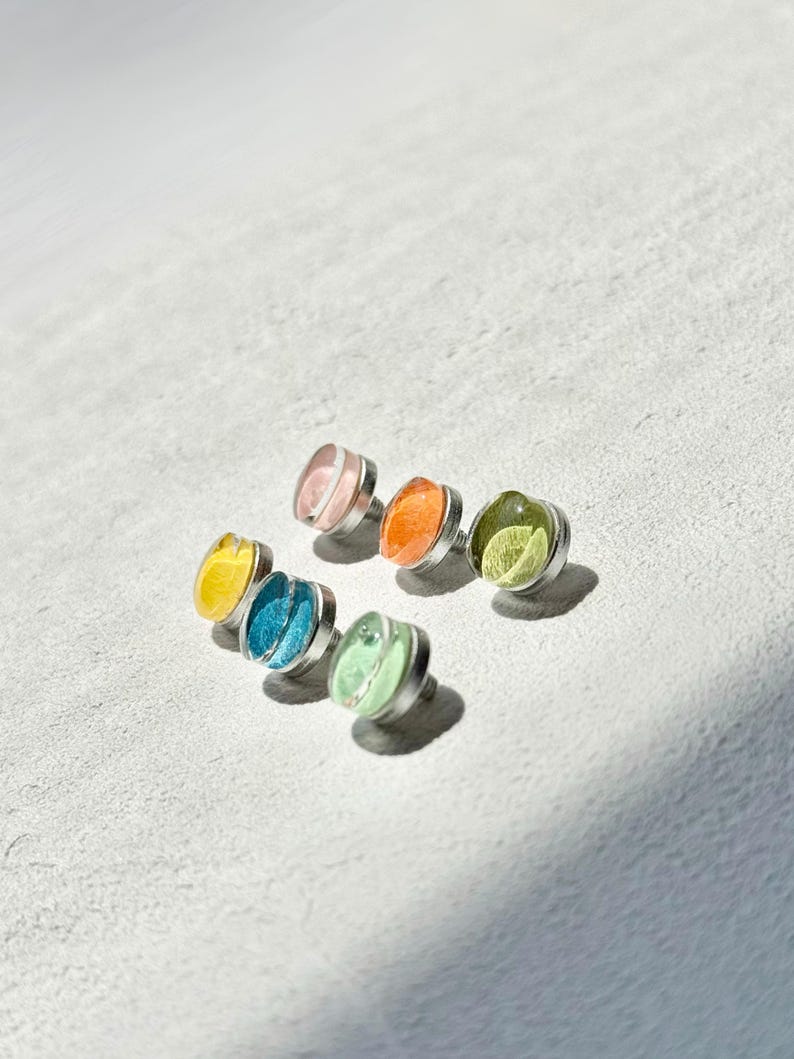 May include: A selection of colourful stud earrings. Each earring has a silver-toned metal base and a translucent, jewel-toned centre in shades of pink, orange, green, blue, and yellow. The earrings are arranged on a light grey surface.