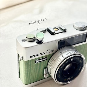 May include: A vintage Konica C35 camera with a mint green and silver finish. The camera features a Hexanon lens and the text "Flashmatic". The words "mint green" are written above the camera.