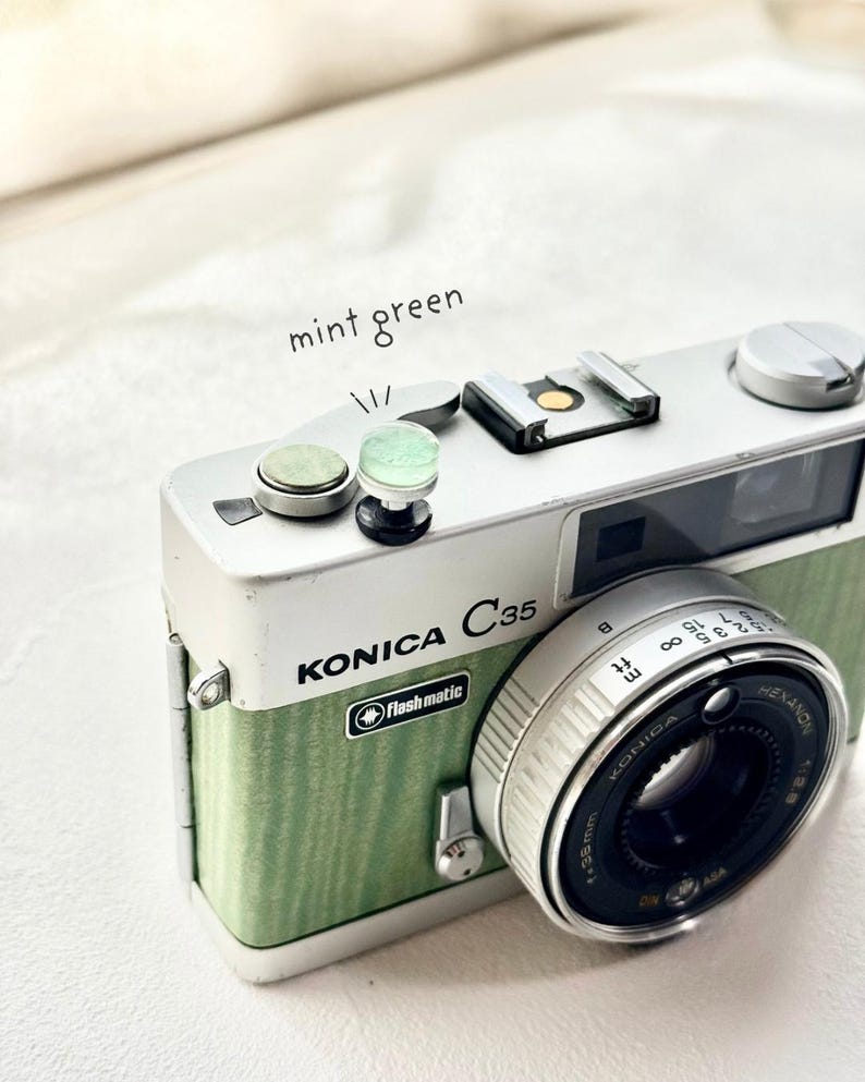 May include: A vintage Konica C35 camera with a mint green and silver finish. The camera features a Hexanon lens and the text "Flashmatic". The words "mint green" are written above the camera.
