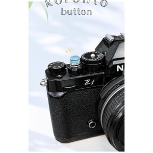 May include: A close-up of a black camera with a textured body and silver details. The camera features a blue button and the text "koronto button" is above the camera. The lens is visible on the right.
