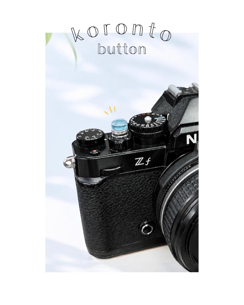 May include: A close-up of a black camera with a textured body and silver details. The camera features a blue button and the text "koronto button" is above the camera. The lens is visible on the right.