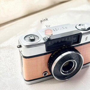May include: A vintage Olympus Pen-EE S camera with a silver body and a light pink leather-like covering. The lens is black, and the camera has a pink button on top. The word "Olympus" is visible on the front.