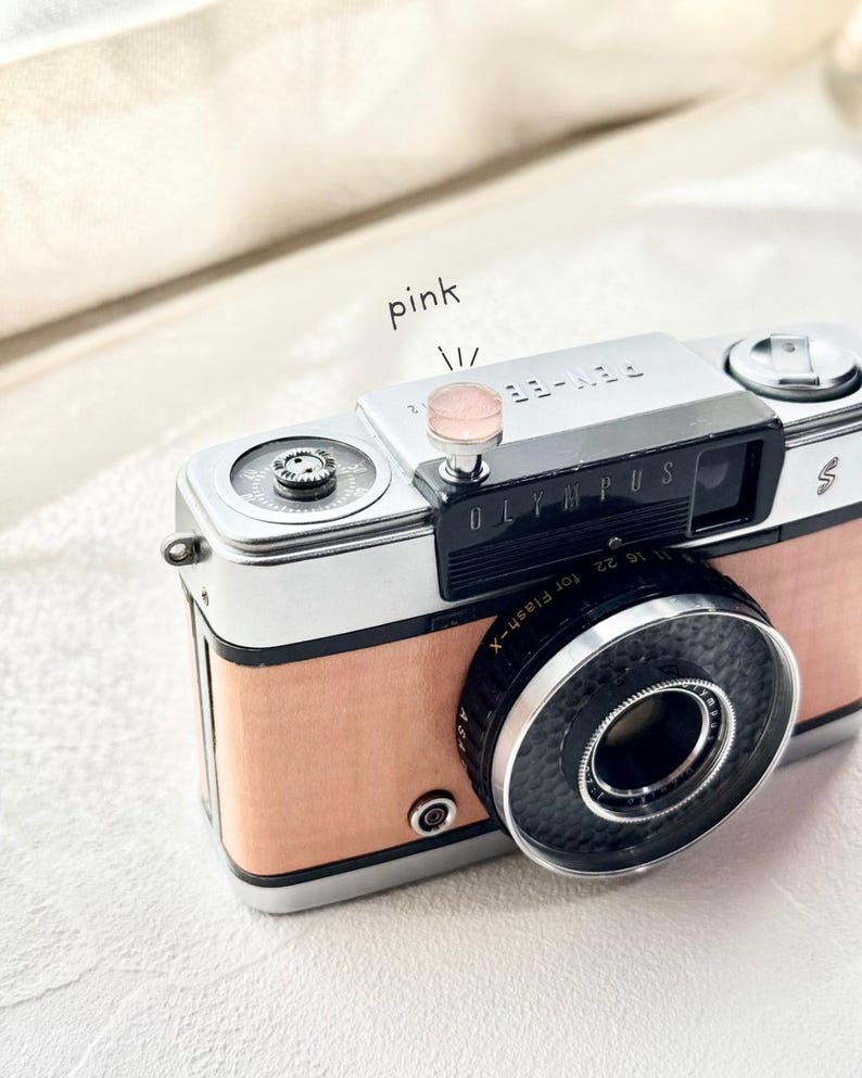 May include: A vintage Olympus Pen-EE S camera with a silver body and a light pink leather-like covering. The lens is black, and the camera has a pink button on top. The word "Olympus" is visible on the front.