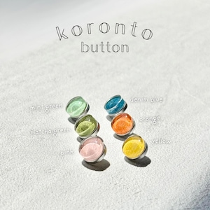 May include: Six colourful buttons with a clear, domed top and silver metal base. The buttons are in shades of mint green, matcha green, pink, denim blue, orange, and yellow. The text "koronto button" is above the buttons.