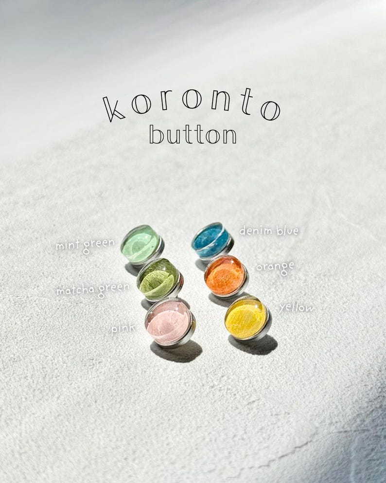 May include: Six colourful buttons with a clear, domed top and silver metal base. The buttons are in shades of mint green, matcha green, pink, denim blue, orange, and yellow. The text "koronto button" is above the buttons.