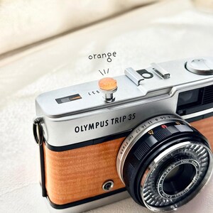 May include: A classic Olympus Trip 35 camera, featuring a silver body and orange wood-effect panelling. The lens is black with silver detailing. The word "orange" is written above the camera's button.