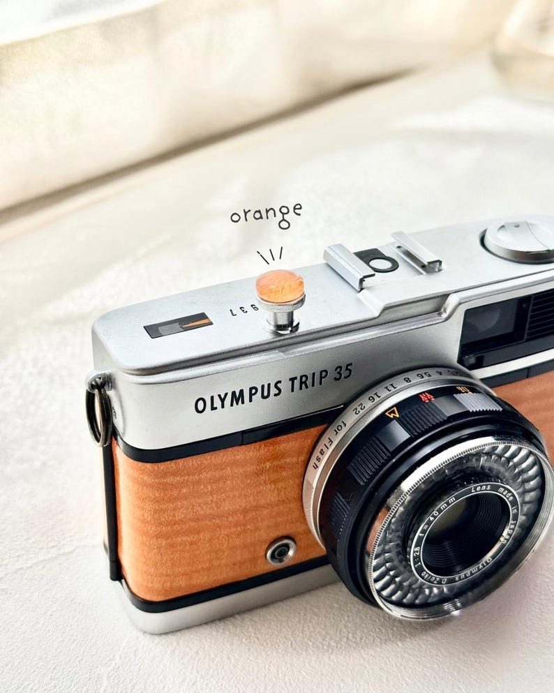 May include: A classic Olympus Trip 35 camera, featuring a silver body and orange wood-effect panelling. The lens is black with silver detailing. The word "orange" is written above the camera's button.